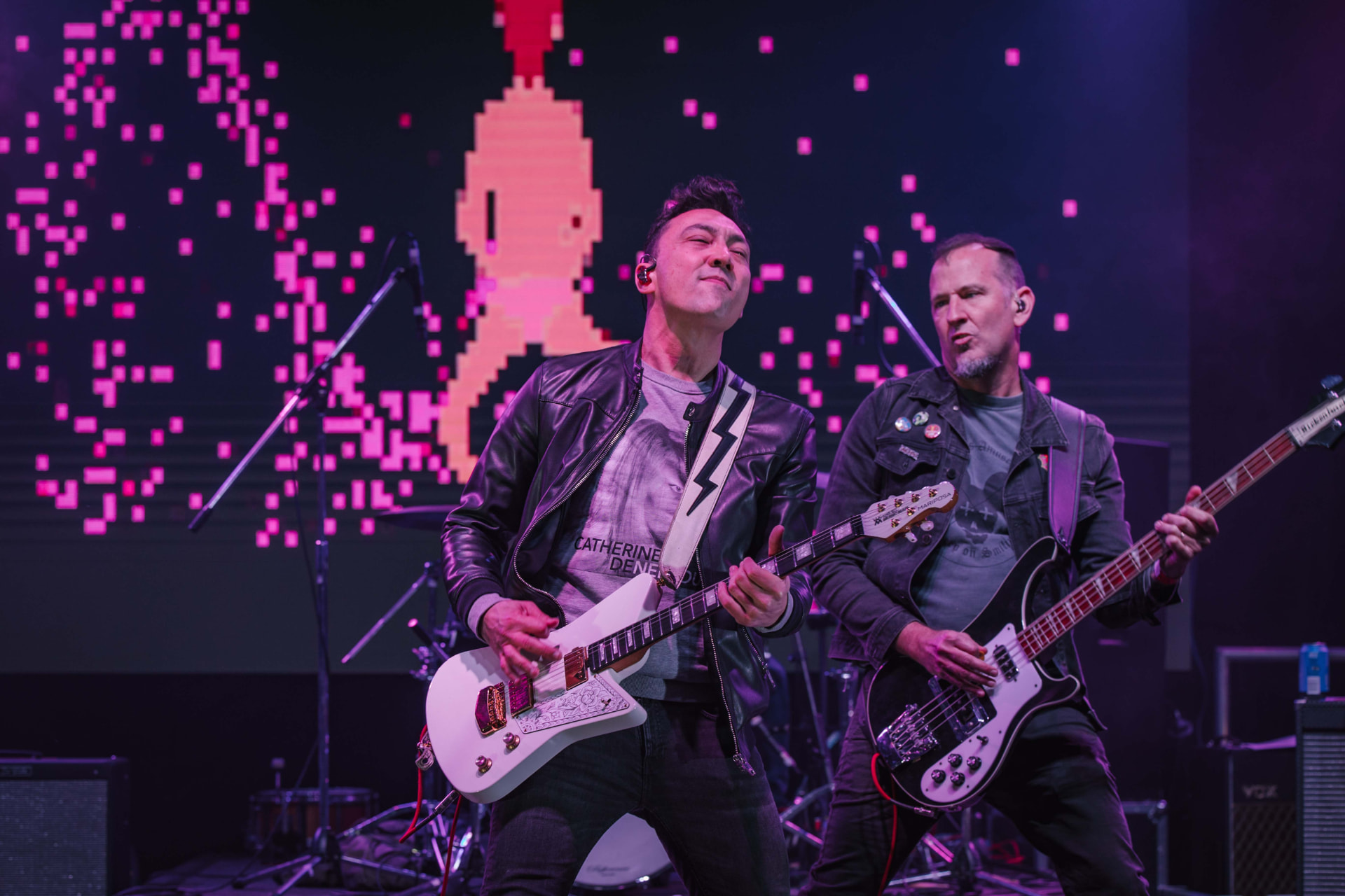 Two musicians performing live on a stage washed in magenta and purple light