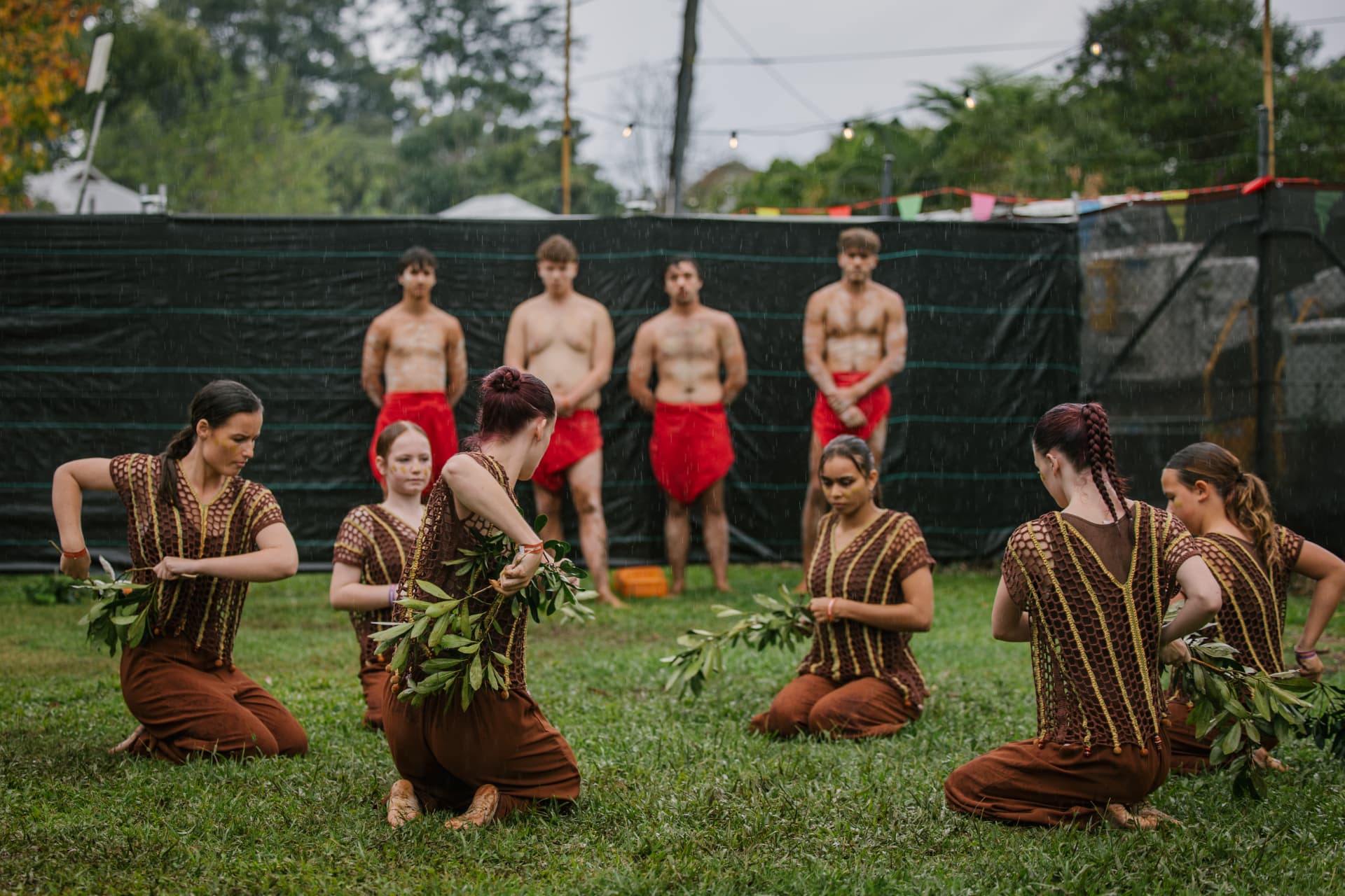 First Nations dancers performing on the grass, kneeling with gum branches while others stand behind