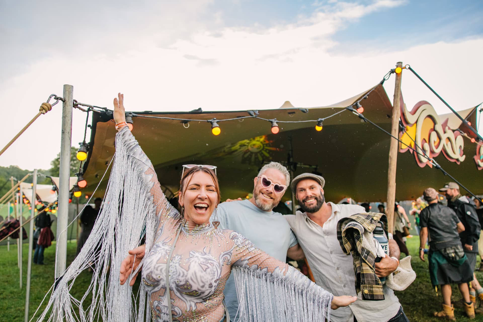 Three festival-goers posing together on the lawn, one in a fringed silver bodysuit with arms outstretched