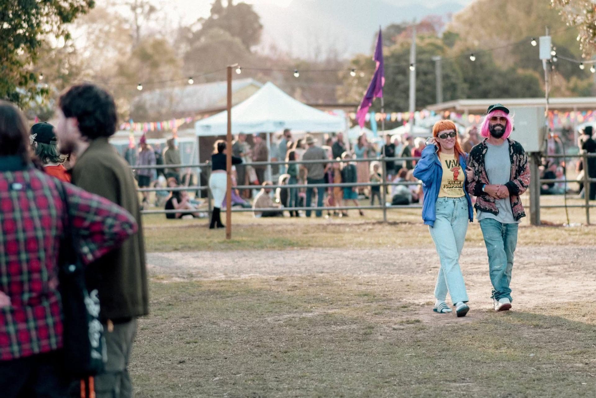 Two festival-goers walking arm in arm through the festival village, one wearing a bright pink wig