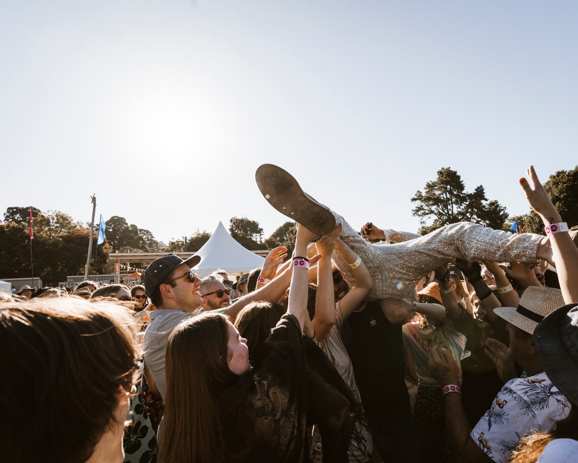 A performer in a silver sequin suit being crowd-surfed above a sunlit crowd